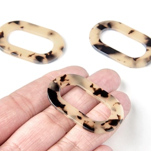 Peut inclure: Trois maillons ovales en acrylique à motif écaille de tortue. Les maillons sont de couleur beige clair avec des taches marron foncé. Un maillon est tenu dans une main, et deux sont sur une surface blanche.