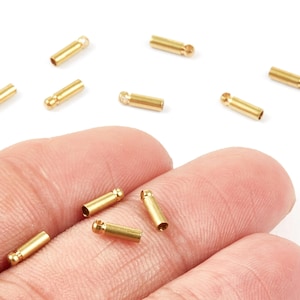 Peut inclure: Gros plan sur de petites perles tubulaires en métal doré, chacune avec une boucle à une extrémité. Plusieurs perles sont éparpillées sur une surface blanche, certaines sont tenues dans une main. Idéales pour la création de bijoux.