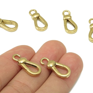 May include: Several small, gold-colored metal clasps with a hook and a spring-loaded closure. Two clasps are held in a hand, while others are scattered on a white surface. These clasps are likely used for jewelry making or crafting.