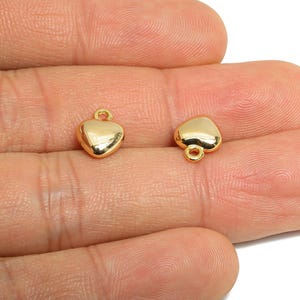 May include: Two small, gold-colored heart-shaped charms. Each charm has a loop at the top for attaching to a chain or other jewelry component. The charms are smooth and shiny, reflecting light.