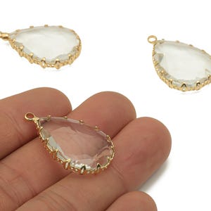 Puede incluir: Colgantes de cristal transparente en forma de lágrima con bordes de metal dorado. Los colgantes tienen un lazo en la parte superior para sujetarlos a una cadena u otro componente de joyería. La imagen muestra tres colgantes, uno de ellos en una mano.