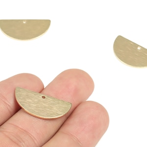 May include: Three gold-colored, half-moon shaped metal charms with a brushed finish. One charm is held in a hand, while two others are isolated against a white background. Each charm has a small hole for stringing.