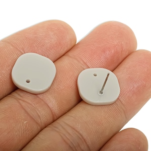 May include: Two light gray, square-shaped stud earrings. One earring has a post and the other has a hole. The earrings are held in a person's hand. The earrings are a neutral color and have a smooth surface.