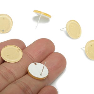 May include: Pale yellow, round stud earrings with silver posts. The earrings have a speckled appearance and a small hole near the edge. Several earrings are visible, some with the backs exposed.