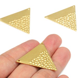 Peut inclure: Trois composants de bijoux dorés en forme de triangle. Chaque composant a une section supérieure lisse et une section inférieure texturée, martelée. Les composants ont de petits trous pour l'attache. Un composant est tenu dans une main.