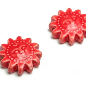 May include: Two red ceramic sun-shaped beads with a raised face design. The beads have a textured edge.