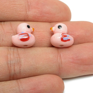 May include: Two small, pink duck beads with black eyes and orange beaks. Each duck has red and blue painted details. The beads are held in a hand, and are likely for jewellery making or crafts.