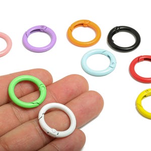May include: A collection of colorful, round spring snap clasps in various shades including pink, purple, orange, black, light blue, red, green, white, and yellow. The clasps are shown on a white background, with some held in a hand.