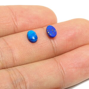 May include: Two small, oval-shaped gemstones in shades of blue are displayed. One is a vibrant, solid blue, while the other has a darker blue base with flecks of gold. The gemstones are likely for jewelry making.