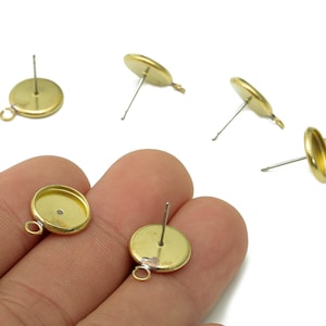 May include: A collection of gold-coloured earring posts. Each post has a flat, circular setting and a small loop. The posts are designed for earrings, with a straight pin for insertion. The settings are intended to hold decorative elements.