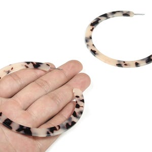May include: A pair of large, open hoop earrings with a tortoiseshell pattern. The earrings are a light beige color with dark brown and black spots. One earring is held in a hand, and the other is separate.
