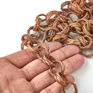 Puede incluir: Primer plano de una cadena trenzada marrón, con una mano sosteniendo una parte de ella. Los eslabones de la cadena están hechos de material marrón claro, tejido apretadamente. El fondo es blanco, resaltando la textura y el color de la cadena.