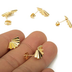 May include: Gold-colored stud earrings with a fan-like design. The earrings have a post and butterfly backing. The image shows several pairs of the earrings on a white background.