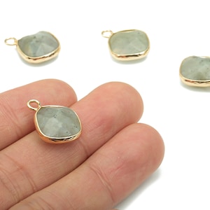 May include: Four square-shaped gemstone pendants with gold-colored bezels. One pendant is held in a hand. The gemstones are a pale green color and have a faceted surface. Each pendant has a small loop for attaching to a chain or other jewelry component.