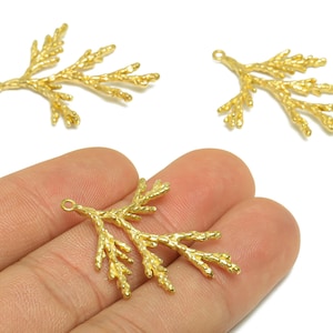 Puede incluir: Encantos de rama de metal de color dorado con una superficie texturizada. Los amuletos tienen un pequeño lazo en la parte superior para sujetarlos a las joyas. Un amuleto se sostiene en una mano, con otros dos sobre un fondo blanco.