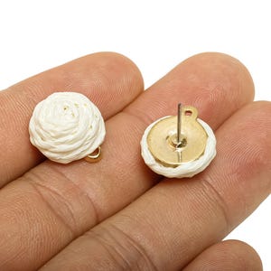 May include: A pair of white, woven-style stud earrings. Each earring features a round, cream-coloured woven design on a gold-coloured base. The earrings are shown on a person's hand.