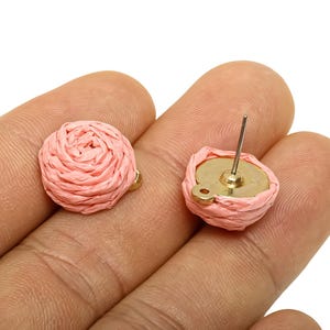 May include: A pair of pink woven stud earrings. The earrings feature a circular design with a woven texture, resembling a rose. The earring posts are silver, and the base is gold-toned.