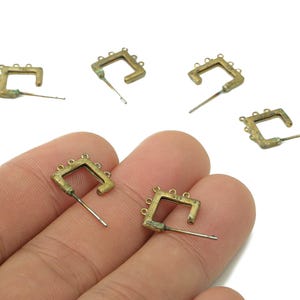 May include: Several small, square-shaped brass earrings with a post and three small loops on each corner are scattered on a white surface. The earrings have a vintage, antique appearance.