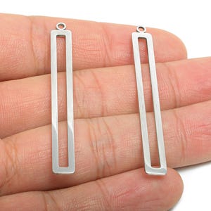 Può includere: Due ciondoli rettangolari argentati con un centro cavo e un piccolo anello nella parte superiore. I ciondoli sono tenuti nella mano di una persona, mostrando il loro design elegante e minimalista. Ideali per la creazione di gioielli.