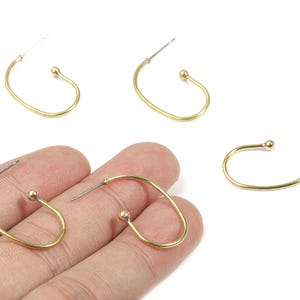 May include: Gold-colored hoop earrings with a small ball accent at the bottom. The earrings have a curved, open design and a post for insertion. Several earrings are displayed on a white background, with one pair held in a hand.