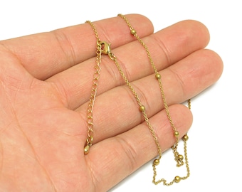 May include: A delicate gold-colored necklace with small, evenly spaced beads. The necklace is held in an open hand against a white background, showcasing the chain's fine details and the clasp.