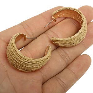 May include: A pair of gold-coloured hoop earrings with a woven, raffia-like texture. The earrings are crescent-shaped and have a post closure. The earrings are displayed on a hand against a white background.