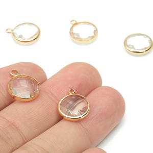 May include: Four clear, round crystal charms with gold-colored metal edging. The charms have a small loop at the top for attaching to jewelry. Two charms are held in a hand, and two are on a white surface.
