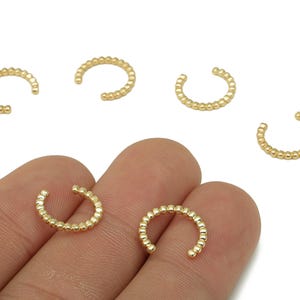 May include: Gold-colored ear cuffs with a beaded design. The open-ended, C-shaped cuffs are shown in a close-up view, with some resting on a hand. The jewelry is set against a white background.