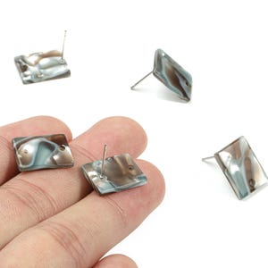 May include: Square stud earrings with a marbled pattern in shades of brown, gray, and blue. The earrings have a post backing. Several earrings are scattered on a white background.