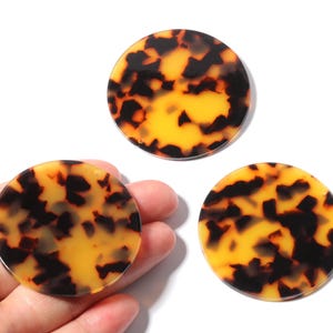 Könnte beinhalten: Drei runde Acrylscheiben mit Schildpattmuster. Die Scheiben weisen eine Mischung aus dunkelbraunen und gelb-orangenen Farbtönen auf, wodurch ein marmorierter Effekt entsteht. Eine Scheibe wird in einer Hand gehalten, die anderen beiden werden separat angezeigt.
