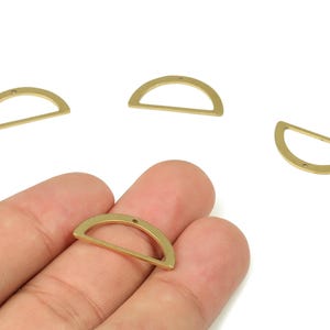 May include: Several brass-colored, semi-circular jewelry findings. One is held in a hand, with others scattered on a white surface. Each has a small hole for attaching to other components.