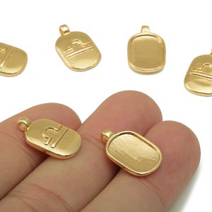 May include: Gold-coloured Libra zodiac sign charms. The charms are rectangular with rounded edges and a small loop at the top. The Libra symbol is embossed on some charms. The charms are shown on a white background.