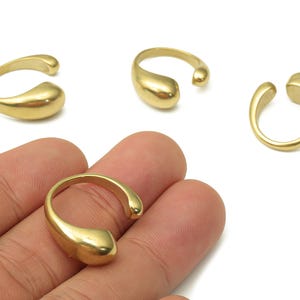 May include: Gold-colored open-ended rings with a teardrop design. The rings are shown on a white background, with one ring being held by a hand. The rings have a smooth, polished finish and a simple, modern style.