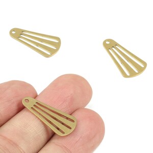Puede incluir: Tres dijes de metal dorado en forma de abanico con un pequeño agujero en la parte superior para ensartar. Cada dije tiene tres líneas paralelas recortadas. Un dije se sostiene en una mano y dos están sobre un fondo blanco.
