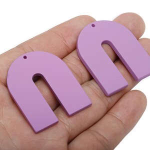 以下が含まれることがあります： 2つのラベンダー色のU字型イヤリング。各イヤリングには、吊り下げるための小さな穴が上部にあります。イヤリングは滑らかで、マットな仕上がりです。イヤリングは人の手で持たれています。