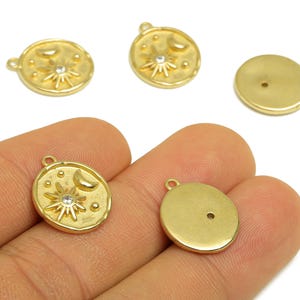 Könnte beinhalten: Goldfarbene Himmelsanhänger mit Sonnen-, Mond- und Sterndesigns. Jeder Anhänger hat eine kleine Schlaufe zum Befestigen an Schmuck. Ein Anhänger hat einen klaren Kristall in der Mitte der Sonne. Die Anhänger sind rund und haben eine strukturierte Oberfläche.