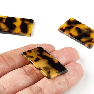 May include: Rectangular tortoise shell beads with a hole at the top for stringing. The beads have a mottled pattern of dark brown and yellow. Three beads are visible, one held in a hand.