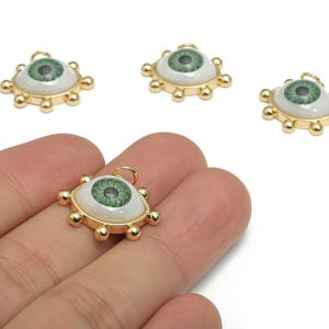 Op de afbeelding: Oogvormige hangerbedels met een goudkleurig frame en een groene iris. De bedels hebben een witte achtergrond en zijn omgeven door goudkleurige kralen. De oogbedels worden op een witte achtergrond getoond.