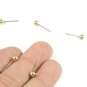 Puede incluir: Primer plano de varios pequeños postes de pendientes dorados en forma de bola con postes plateados. Los pendientes están sobre un fondo blanco. Un pendiente se sostiene entre los dedos.