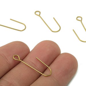 Puede incluir: Primer plano de varios ganchos de pendientes dorados. Un gancho se sostiene entre los dedos, mientras que otros tres están dispersos sobre un fondo blanco. Los ganchos tienen un diseño simple y minimalista, con una parte superior en bucle y una parte inferior curvada.