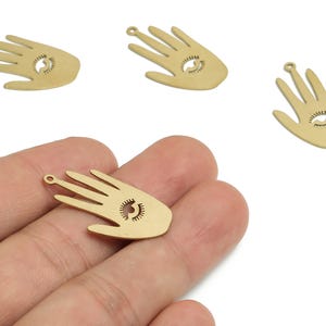 Może przedstawiać: Złote metalowe zawieszki w kształcie dłoni z motywem oka. Zawieszki mają wycięte oko na dłoni i małą pętelkę u góry do mocowania do biżuterii. Zawieszki są prawdopodobnie przeznaczone do tworzenia biżuterii DIY.