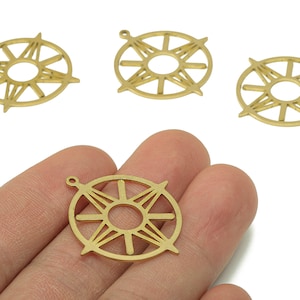 May include: Four gold-coloured compass charm pendants. One pendant is held in a hand, while three others are scattered nearby. Each charm features a circular design with directional lines and a small loop for attaching to jewellery.