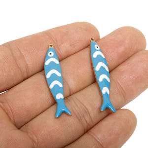 Puede incluir: Dos colgantes azules con forma de pez con patrones de chevrón blancos y lazos dorados. Los colgantes están esmaltados y tienen un pequeño detalle de ojo. Los colgantes de pez son un elemento decorativo para la fabricación de joyas.
