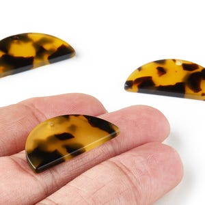 May include: Three half-circle shaped pendants with a tortoiseshell pattern. The pendants are a mix of yellow and dark brown, with a glossy finish. One pendant is held in a hand.