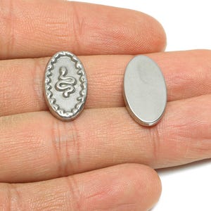 Puede incluir: Dos colgantes plateados de forma ovalada. Uno presenta un diseño de serpiente dentro de un borde decorativo. El otro colgante es un óvalo liso. Estos colgantes son probablemente para la fabricación de joyas o manualidades.