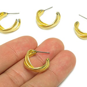 Peut inclure: Boucles d'oreilles créoles dorées avec un design torsadé. Les boucles d'oreilles sont petites, avec une tige pour l'insertion dans l'oreille. Plusieurs boucles d'oreilles sont visibles, certaines sur une surface blanche et une tenue dans une main.