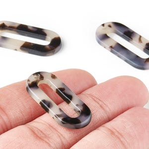 Può includere: Tre perline ovali in resina con motivo tartarugato. Le perline sono un mix di crema, nero e marrone. Una perlina è tenuta in mano, mentre le altre due sono su una superficie bianca.