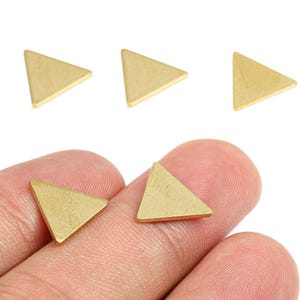 May include: Close-up of several small, gold-colored triangle charms. Two charms are held between fingers, while three others are scattered above. The charms have a matte finish and are likely for jewelry making or crafting.