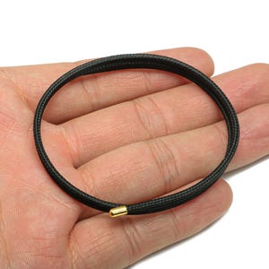 Può includere: Un braccialetto nero con una trama intrecciata e una chiusura color oro. Il braccialetto è circolare e sembra essere realizzato in un materiale resistente. Il design è semplice ed elegante, adatto all'uso quotidiano.