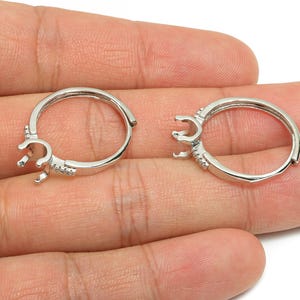 May include: Two silver-colored hoop earring findings with a prong setting for gemstones. The earrings are open, showing the clasp mechanism. Small, clear stones are set along the hoop. The earrings are held in a hand.